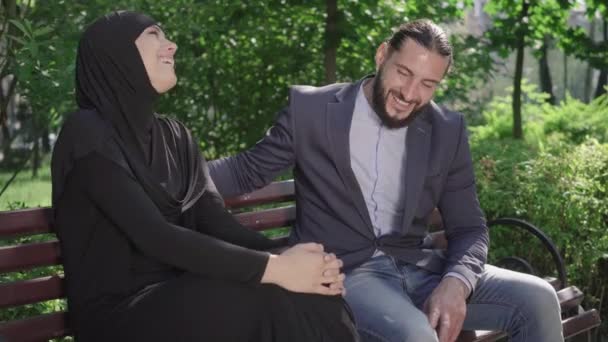 幸せな笑いのイスラム教徒のカップル公園でベンチに座っておしゃべり。屋外で晴れた夏の日を楽しむヒジャーブ州の喜びの愛する髭の男と女の肖像画。デート,愛,ロマンス,ライフスタイル. — ストック動画