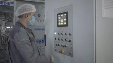 Yüz maskeli bir adamın fabrikadaki endüstriyel ekipmanları açtığının yan görüntüsü. Beyaz erkek çalışan otomatik makinelerin düğmelerine basıyor ve yeşil diyotlar yanıyor. Endüstri, üretim.