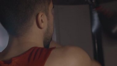 Kafkas boksörünün siyah spor salonunda kum torbasına vuruşunu yakından çek. Esmer bir sporcunun teçhizatıyla omuz üstünden atış yapmak. Güç, dövüş sanatları, dövüş sporları. Sinema 4k ProRes Merkezi.