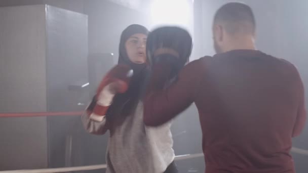 Serious concentrated muslim female boxer punching training gloves. Young woman in hijab exercising with coach in haze with backlight at the background. Sport, gender equality. Cinema 4k ProRes HQ. — Stock Video