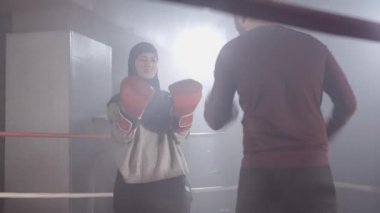 Pozitif Müslüman kadın ve beyaz adam boks ringinde sisli bir şekilde antrenman yapıyor. Türbanlı genç sporcunun idmanı ve arka planda kişisel antrenör. Dövüş sporu, yaşam tarzı. Sinema 4k ProRes Merkezi.