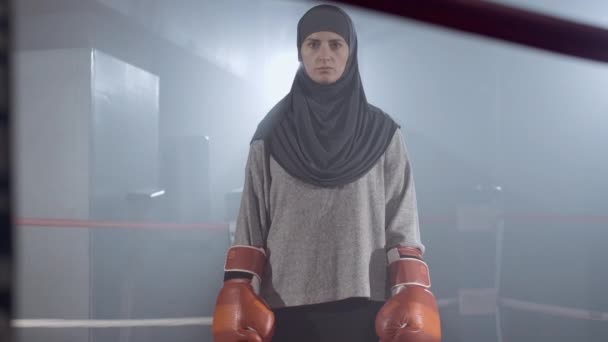 Portrait de femme musulmane sérieuse en hijab levant la main dans des gants de boxe et regardant la caméra. boxeuse forte confiante debout sur anneau brumeux avec rétro-éclairage. Confiance. Siège social Cinema 4k ProRes. — Video