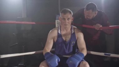 Beyaz tenli terli bir boksörün portresi köşede oturuyor ve koç onu destekliyor. Atletik, kendine güvenen genç sporcu arka plandaki boks ringinde dinleniyor. Spor, dövüş sanatları..