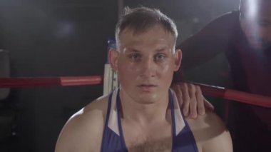 Boks ringinin köşesinde oturan terleyen genç boksörü destekleyen antrenör. Kamera, Kafkasyalı sporcunun savaşa hazır kızgın yüzüne yaklaşıyor. Dövüş sporu, rekabet, şampiyonluk.