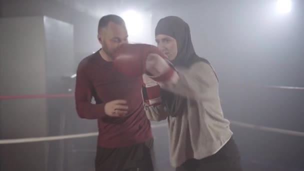 Coach kijavítja a fiatal, gyönyörű muszlim nő boksz pozícióját hidzsáb kiképzésen a ködben a háttérben. Portré magabiztos női bokszoló gyakorló személyi edző lencse fáklya és köd. — Stock videók