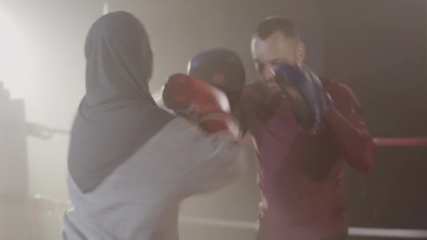 Achteraanzicht van jonge vrouw in hijab ponsen trainingshandschoenen van coach in achtergrondverlichting mist. Zelfverzekerde moslim vrouwelijke bokser training op boksring in lense flare. Bestrijding van sport, gendergelijkheid. — Stockvideo