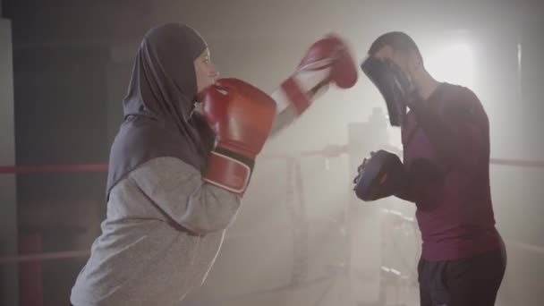 侧视图的穆斯林女子拳击手冲压训练手套作为教练说话.集中的年轻女运动员与私人教练在薄雾中的镜片照明弹中在拳击台上战斗的肖像。体育. — 图库视频影像