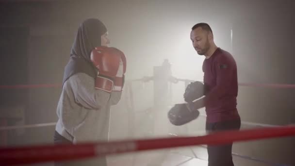 Trening muzułmańskich sportowców w hidżabie na ringu bokserskim. Widok z boku portret pewny siebie trener korygujący postawę bokserki w mgle w podświetleniu. Sparing mężczyzny i kobiety we mgle rozbłysku soczewki. — Wideo stockowe