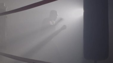 Arka planda ışıkla sisli havayı yumruklayan boksör silueti. Kafkasyalı sporcuların sisli spor salonunda boks ringinde yaptıkları antrenmanların geniş açılı görüntüsü. Sinema 4k ProRes Merkezi.