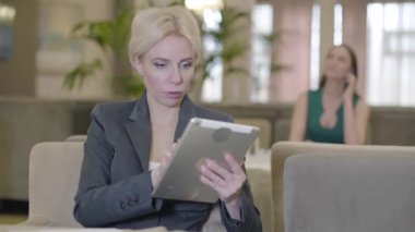 Restoranda sosyal medyayı tablette kullanan heyecanlı sarışın kadının portresi. İnternet 'te sörf yapan ve gülümseyen beyaz takım elbiseli güzel bir bayan. Arka planda telefonla konuşan bulanık kız..