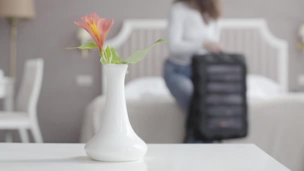 Close-up de flor em vaso em pé à mesa branca com a mulher borrada sentada na cama e abertura da mala no fundo. Jovem turista caucasiano irreconhecível entrando em acomodações no resort . — Vídeo de Stock