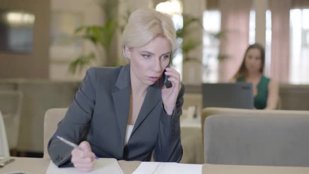 Occupata donna d'affari sicura di parlare al telefono al ristorante e scrivere appunti. Ritratto di donna caucasica stressata con capelli biondi che si occupa di problemi di affari nel caffè. Sovraccarico. — Video Stock