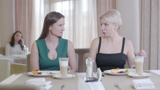 Amigos femininos positivos conversando e comendo sobremesa no restaurante. Retrato de duas jovens mulheres descansando no café. Loira e morena senhoras elegantes degustação de comida doce. Lazer, estilo de vida, amizade, alegria — Vídeo de Stock