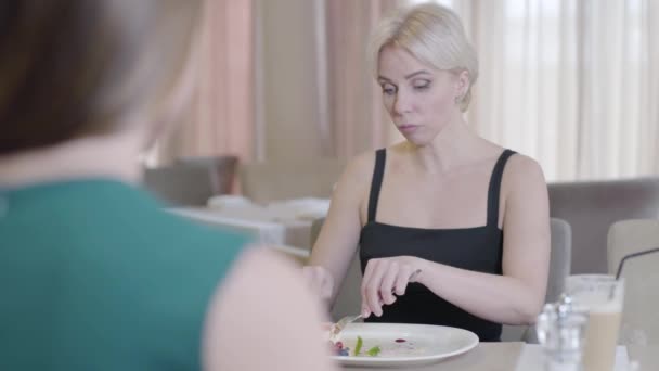 Mujer rubia segura en vestido negro hablando y comiendo sabroso postre en la cafetería. Elegante hermosa dama caucásica charlando con un irreconocible amigo morena en el restaurante. Estilo de vida, comida, ocio . — Vídeo de stock