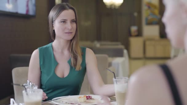 Slim brunette Caucasian woman in green dress talking with unrecognizable friend in cafe. Portrait of beautiful brown-eyed lady listening to blond girl with interest. Communication, friendship. — Stock Video