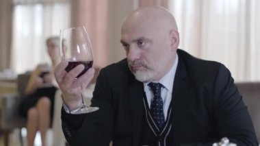 Restoranda kırmızı şarap içen kel, kendine güvenen beyaz adamın yakın plan portresi. Arka planda garson çağıran bulanık sarışın kadın. Takım elbiseli yakışıklı işadamı kafede dinleniyor..