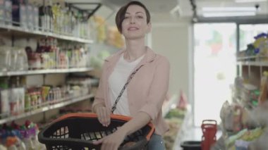 Markette elinde alışveriş sepetiyle poz veren gülümseyen beyaz kadın portresi. Gıda pazarında durup kameraya bakan güzel esmer kadın. Yaşam tarzı, tüketim, ticaret.