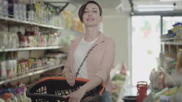 Portret van een glimlachende jonge blanke vrouw die poseert met een boodschappenmandje in de supermarkt. Mooie brunette dame die op de voedselmarkt staat en naar de camera kijkt. Lifestyle, consumentisme, handel. — Stockvideo