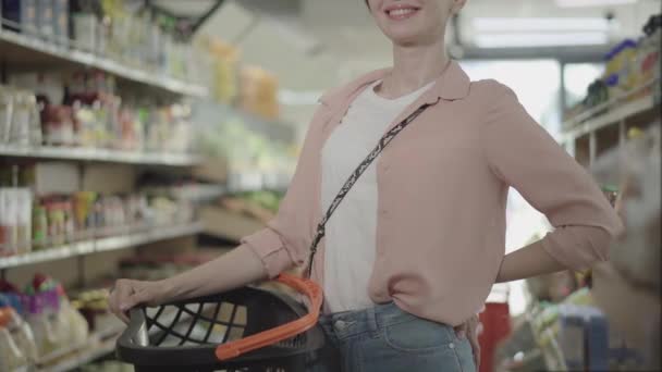 Oigenkännlig leende vit kvinna poserar med shoppingkorg i mataffären. Ung vacker brunett dam stående i matmarknaden. Livsstil, konsumtion, köp, inköp. — Stockvideo