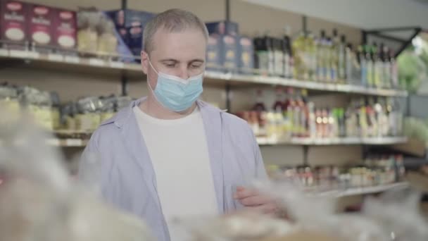 Retrato de homem caucasiano na máscara facial escolhendo produtos no supermercado. Adulto concentrado comprador masculino selecionando bens no mercado de alimentos em quarentena Covid-19. Estilo de vida Coronavirus, compra . — Vídeo de Stock