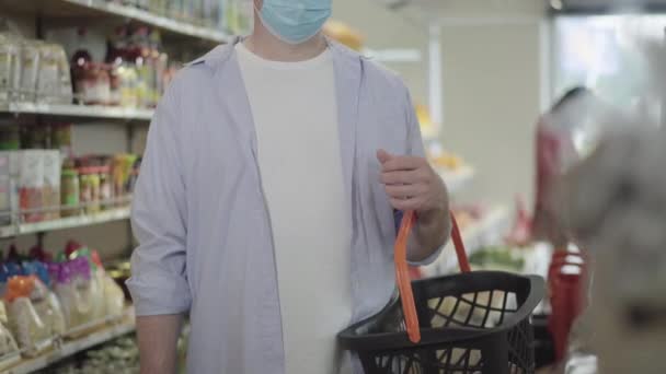 Homem irreconhecível na máscara facial em pé no supermercado com cesta de compras. Adulto branco comprador do sexo masculino comprando bom em Covid-19 pandemia na loja de alimentos. Estilo de vida do coronavírus . — Vídeo de Stock