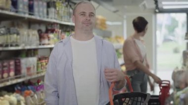 Bakkalda alışveriş sepetiyle poz veren olumlu erkek alıcı arka planda malları seçen bulanık bir kadın olarak görülüyor. Yemek dükkanında durup kameraya gülümseyen mutlu beyaz adamın portresi..