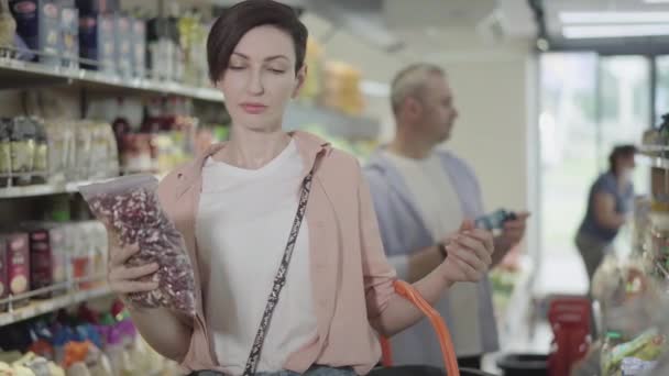 Mujer joven enfocada poniendo envases de frijoles crudos en la cesta de la compra. Hombre borroso elegir productos en el fondo. Abarrotes, mercado de alimentos, compras, compras . — Vídeos de Stock