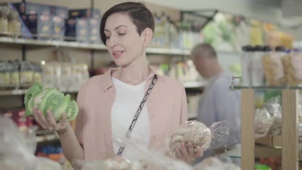 Mooie jonge vrouw kiezen tussen kool en peperkoek koekjes. Portret van brunette dame met bruine ogen selecteren van gezonde en ongezonde producten in de supermarkt. Voedselmarkt, levensstijl. — Stockvideo