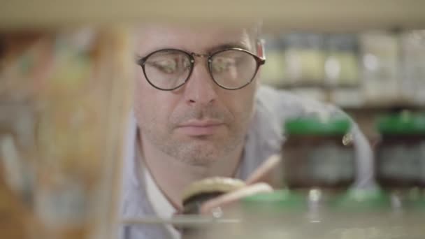 Close-up of man in eyeglasses looking at canned food in grocery and leaving. Portrait of concentrated confident Caucasian buyer choosing products in food market. Shooting behind shelves. — Stock Video