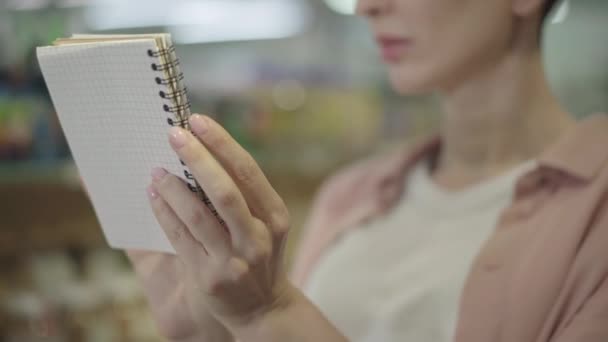 Close-up de mãos caucasianas femininas segurando caderno. Mulher irreconhecível lendo lista de lojas no supermercado. Consumismo, planejamento, estilo de vida, compra . — Vídeo de Stock