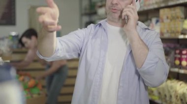 Sıkılmış Kafkasyalı koca, telefonda eşiyle konuşurken marketten ürün seçerken görülüyor. Yetişkin bir adam süpermarkette el kol hareketi yapıyor ve alışveriş yapıyor. Tüketim, yaşam tarzı.