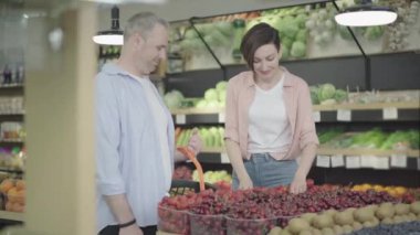 Gülümseyen güzel bir kadın alışveriş sepetine bir sepet lezzetli çilek koyuyor. Beyaz bir adam markette genç ve güzel bir eşle alışveriş yapıyor. Yaşam tarzı, mutluluk..