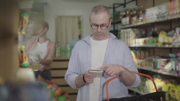 Důvěryhodný muž v brýlích kontroluje seznam obchodů a dává rajčatovou pomazánku do nákupního košíku. Koncentrovaná dospělá běloška kupující zboží v potravinách. Životní styl, nakupování, konzumace. — Stock video