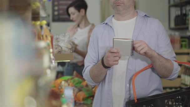 Homem adulto concentrado com lista de compras comprando mercadorias no supermercado. Comprador caucasiano irreconhecível em óculos comprando produtos na loja de alimentos. Consumismo, estilo de vida, concentração . — Vídeo de Stock