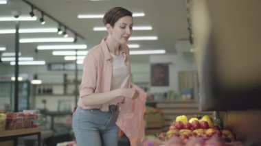 Kendine güvenen genç ev hanımı markette sarı domates seçiyor. Naylon poşete sebze koyan konsantre beyaz kadın portresi. Tüketim, yaşam tarzı, diyet.