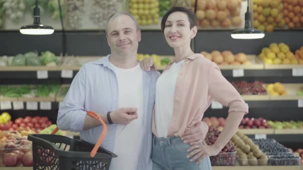 Kaukasische Eheleute umarmen sich und lächeln in die Kamera im Lebensmittelgeschäft. Porträt einer glücklichen jungen schönen brünetten Frau und eines gutaussehenden selbstbewussten erwachsenen Mannes, der im Supermarkt posiert. Lebensstil. — Stockvideo