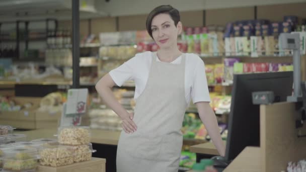 Joven hermosa trabajadora de pie en la caja registradora y sonriendo a la cámara. Retrato de una encantadora morena caucásica con ojos marrones posando en una tienda de comida. Industria, estilo de vida, comercio . — Vídeo de stock