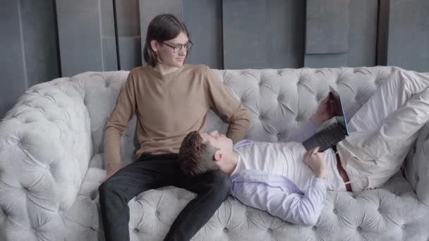 Молодой позитивный мужчина лежит на диване с ноутбуком и разговаривает с партнером-мужчиной. Белая гей-пара отдыхает в помещении по выходным. Любовь, отдых, долгий образ жизни . — стоковое видео