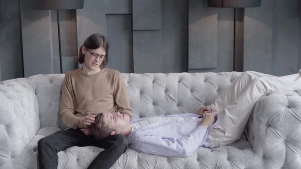 Gelukkig liefhebbende man strelen haar van vriendje liggend op zijn knieën. Portret van een jong blank homoseksueel stel dat geniet van tijd samen thuis en praten. Romantiek, liefde, trots, levensstijl. — Stockvideo