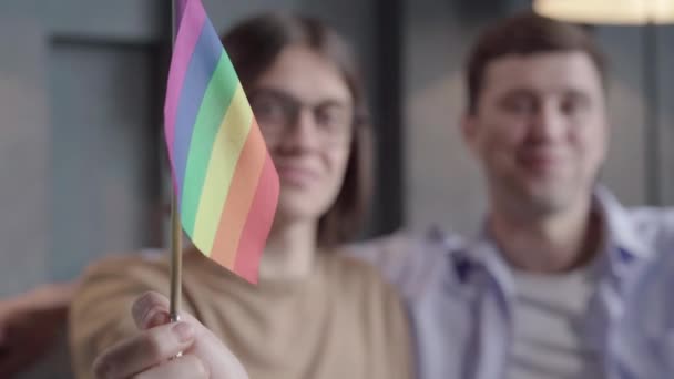 Close-up da bandeira lgbt arco-íris na mão masculina com dois homens borrados sorrindo para o fundo. positivo caucasiano gay casal posando com símbolo dentro de casa. Igualdade, tolerância, estilo de vida, diversidade . — Vídeo de Stock