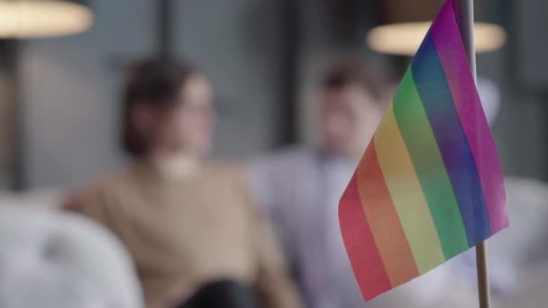 Focus cambia da bandiera lgbt arcobaleno a coppia gay in chat sullo sfondo. Ritratto di uomini caucasici felici e sicuri seduti sul divano a parlare. Simbolo dell'amore dello stesso sesso. — Video Stock