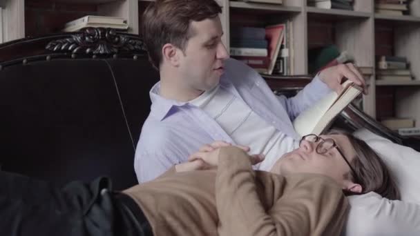 Koncentrerad brunett man läser för gay partner i biblioteket. Porträtt av älskande vit kille liggande på knä av stilig ung make och lyssna på historien. Livsstil, lgbt, hobby, fritid. — Stockvideo