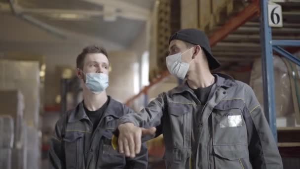 Två vita män i ansiktsmasker diskuterar arbetsplanen på lagret. Porträtt av professionella manliga anställda som arbetar på fabriksförråd på Covid-19 pandemisk karantän. Coronavirus, logistik. — Stockvideo