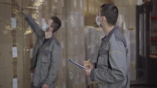 Hombres profesionales en máscaras faciales comprobando la producción en el almacén. Dos empleados caucásicos trabajando en la fábrica. Almacenamiento, fabricación, logística . — Vídeo de stock