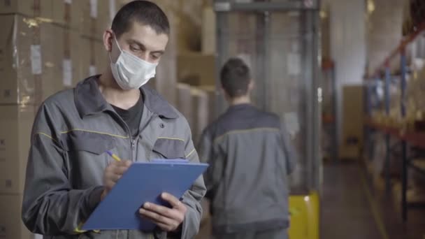 Portrait of brunette man in face mask filling out bill of landing in warehouse. Professional Caucasian male employee working on Covid-19 pandemic quarantine. Coronavirus lifestyle, logistics. — Stock Video