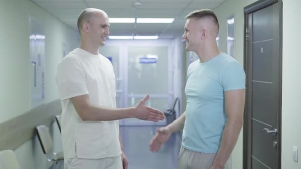 Tevreden cliënt en dokter die elkaar de hand schudden in de ziekenhuisgang en naar de camera kijken. Portret van twee lachende mannen die poseren in de medische kliniek. Begrip gezondheidszorg, geneeskunde, behandeling. — Stockvideo