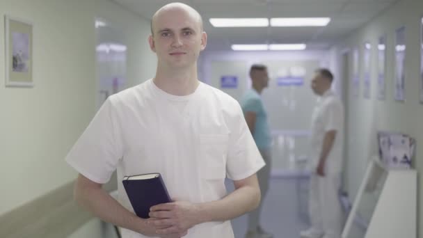 Porträtt av leende ung läkare poserar i sjukhuskorridoren med två suddiga människor skakar hand i bakgrunden. Positiv professionell kaukasisk man som arbetar på läkarkliniken. Hälso- och sjukvård — Stockvideo