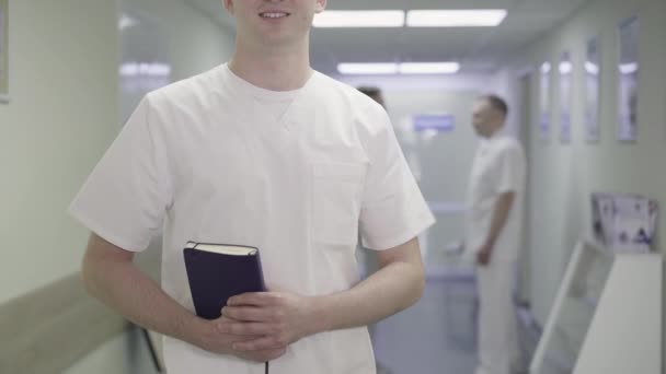 Medico adulto irriconoscibile che posa nel corridoio della clinica medica. Fiducioso uomo caucasico sorridente in piedi sul posto di lavoro in ospedale con uomini sfocati che parlano sullo sfondo. Stile di vita, medicina. — Video Stock