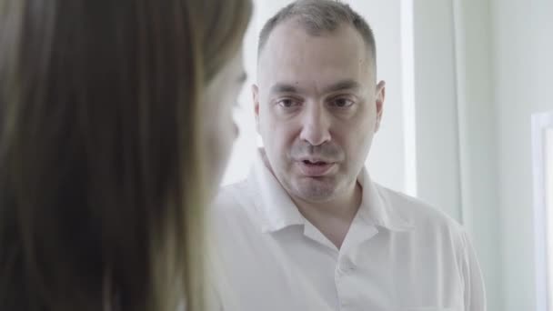 Close-up portrait of Caucasian male doctor consulting young woman in hospital. Confident serious man in white medical robe talking with ill female patient. Health care concept. — Stock Video