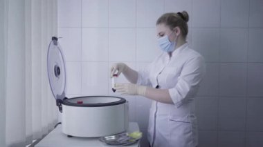 Yüz maskeli ve tıbbi cüppeli kendine güvenen kadın santrifüjden işlenmiş kanla dolu şişeyi çıkarıyor. Covid-19 salgınının laboratuarında çalışan beyaz doktor ya da hemşirenin yan görüntüsü.